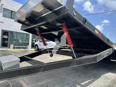 Trailer bed with hydraulic scissor jacks and tilt mechanism in a lowered position, black metal frame, diamond plate toolbox visible, outdoor lot.