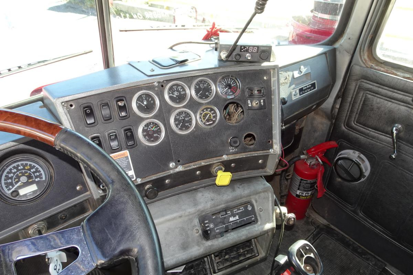 Dashboard of a Mack RD RD688S truck with multiple gauges and switches, steering wheel on left, fire extinguisher mounted on the passenger side, dusty interior.
