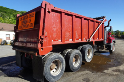 Dump truck rear view showing red dump body with three axles and tires, warning sign on tailgate, water on ground.