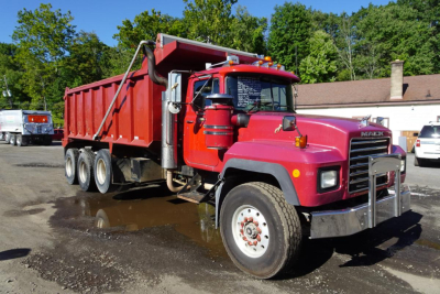 Dump truck chassis and cargo bed visible with red cab and red dump body. Mack RD68S with three rear axles, front grille, and side mirrors.
