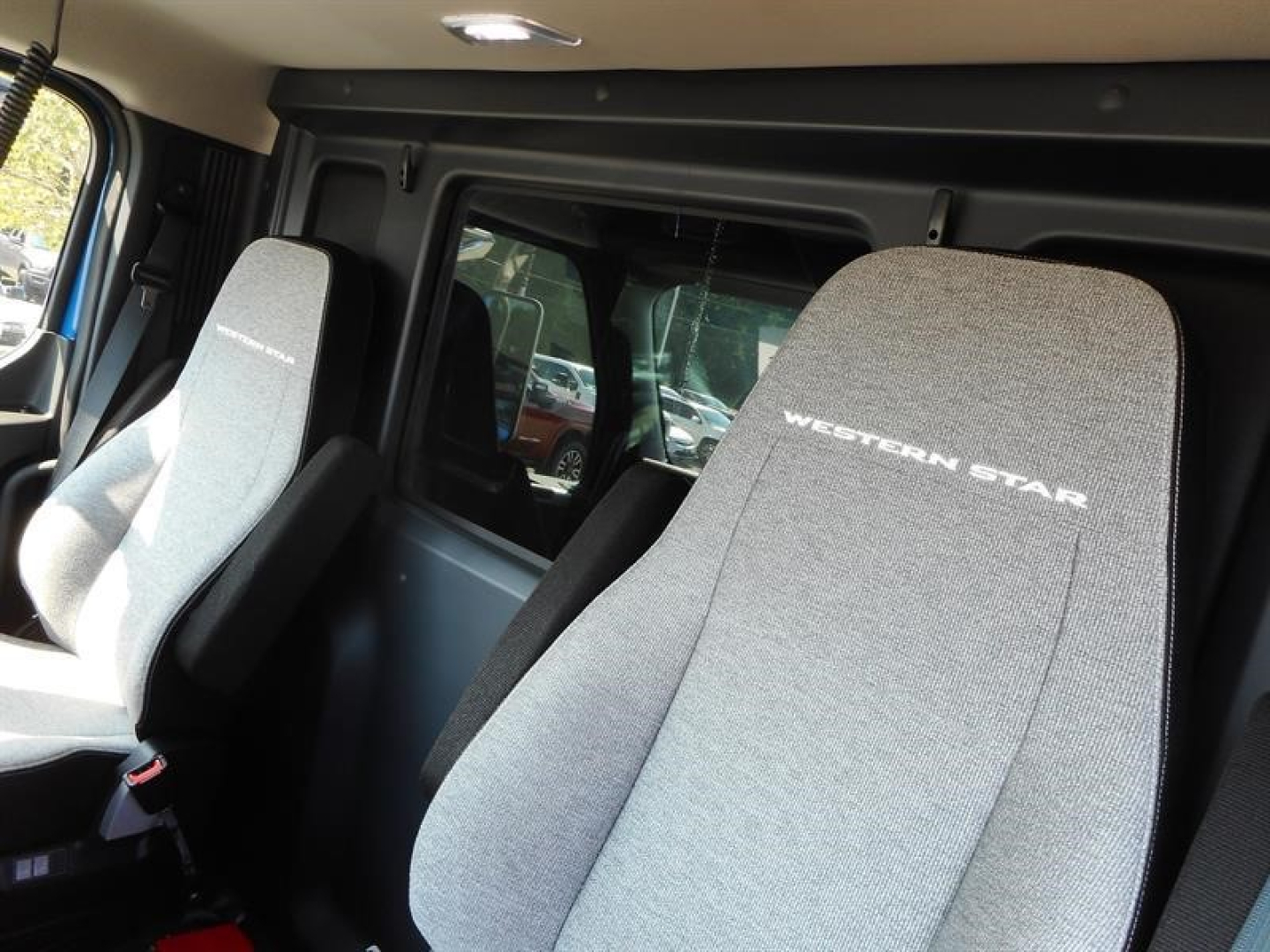 Cab interior of a 2027 Western Star 47X showing driver and passenger seats with light gray fabric and Western Star logo, window behind.