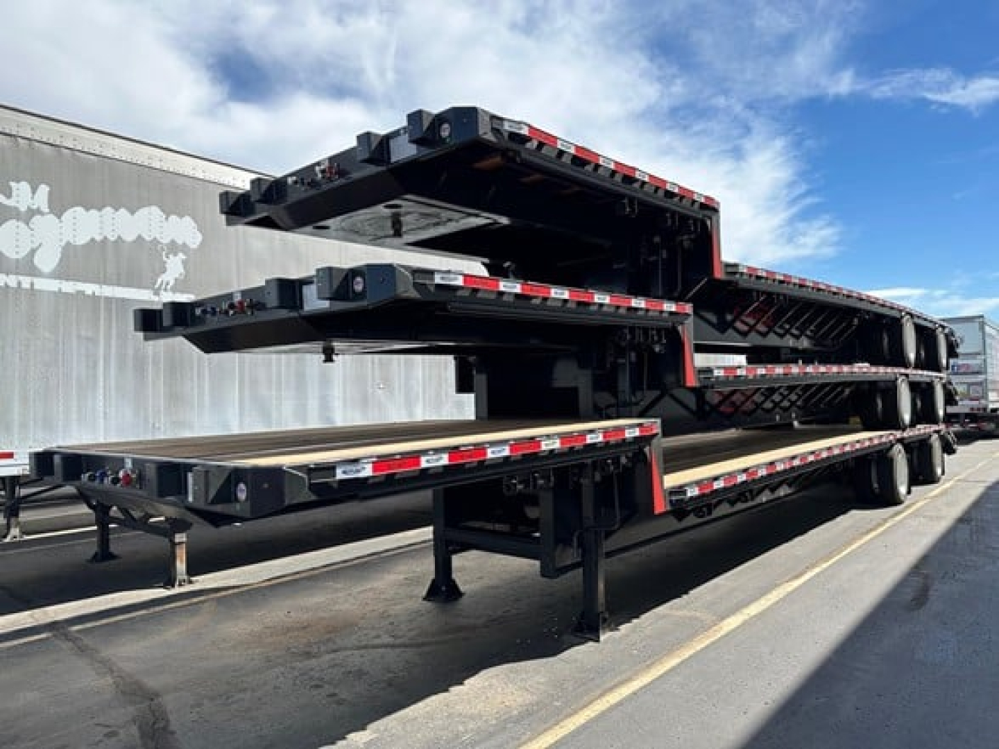 Flatbed trailer with multiple stacked beavertail sections and a long steel trailer frame, showing black paint, red/white reflective tape along edges, and landing gear.