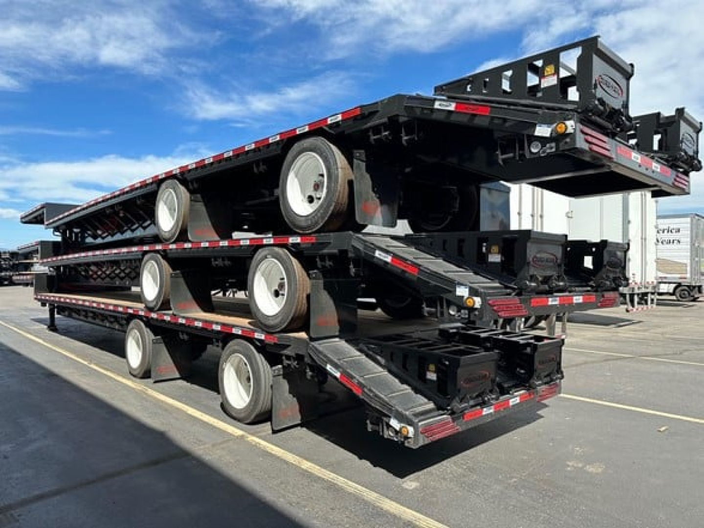 Drop deck trailer with three stacked beavertail-style decks, black metal frame, white wheels, reflective striping, and folded loading ramps.