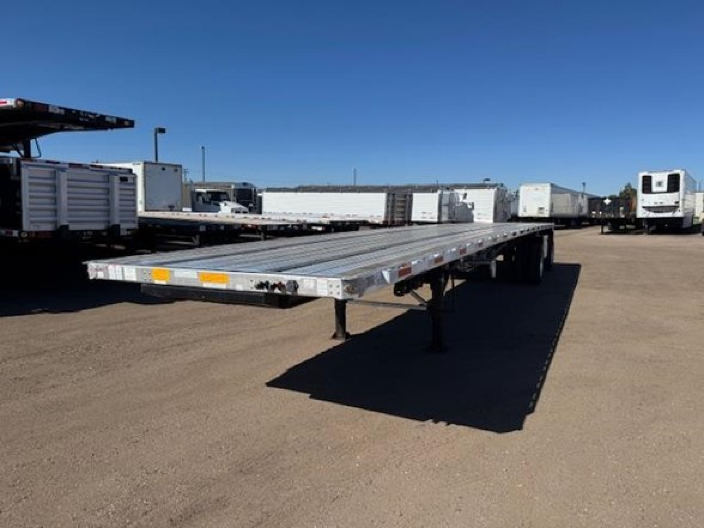 Utility flatbed trailer showing long aluminum deck with dual axles and landing gear, parked in an open lot with other trailers in background.