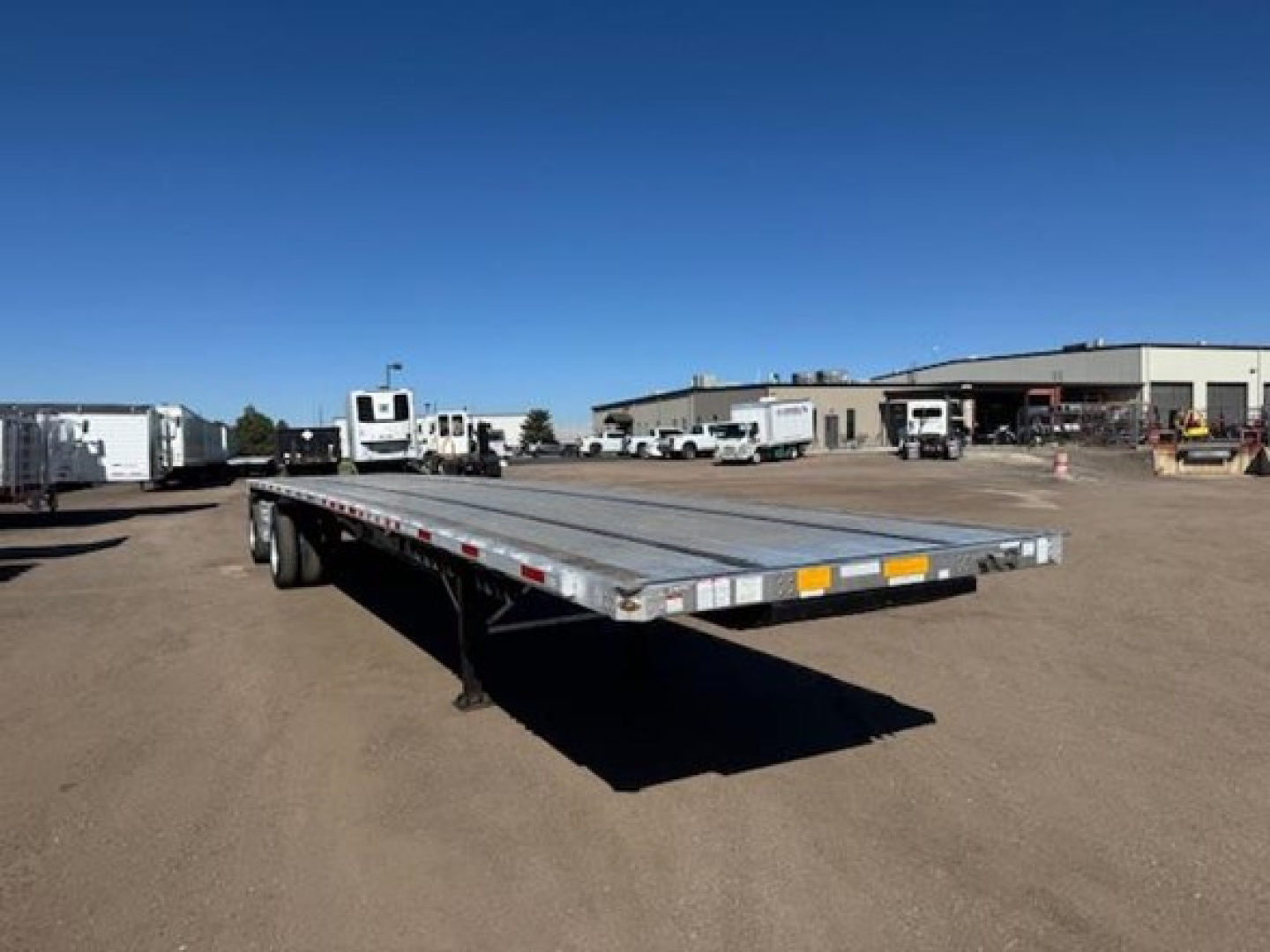 Flatbed trailer in a dealership lot showing a long aluminum deck and dual rear axles under a blue sky.