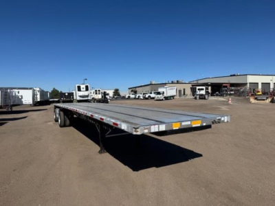 Flatbed trailer in a dealership lot showing a long aluminum deck and dual rear axles under a blue sky.