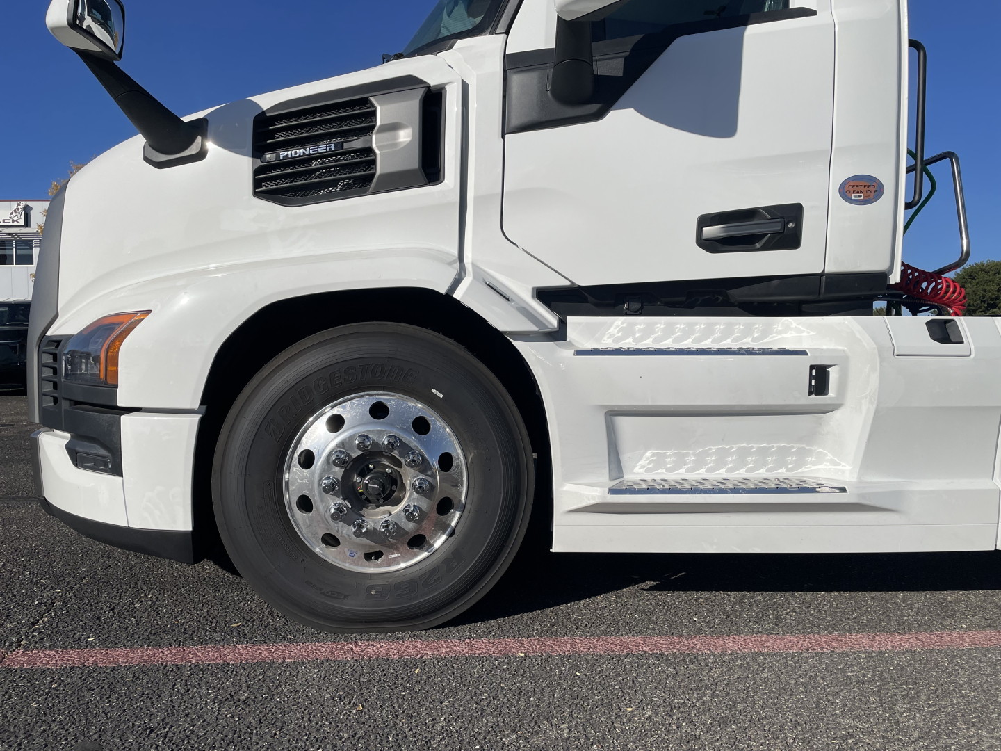 Front-side view of a white 2026 Mack PR64T conventional daycab truck showing front wheel, fender, and steps.