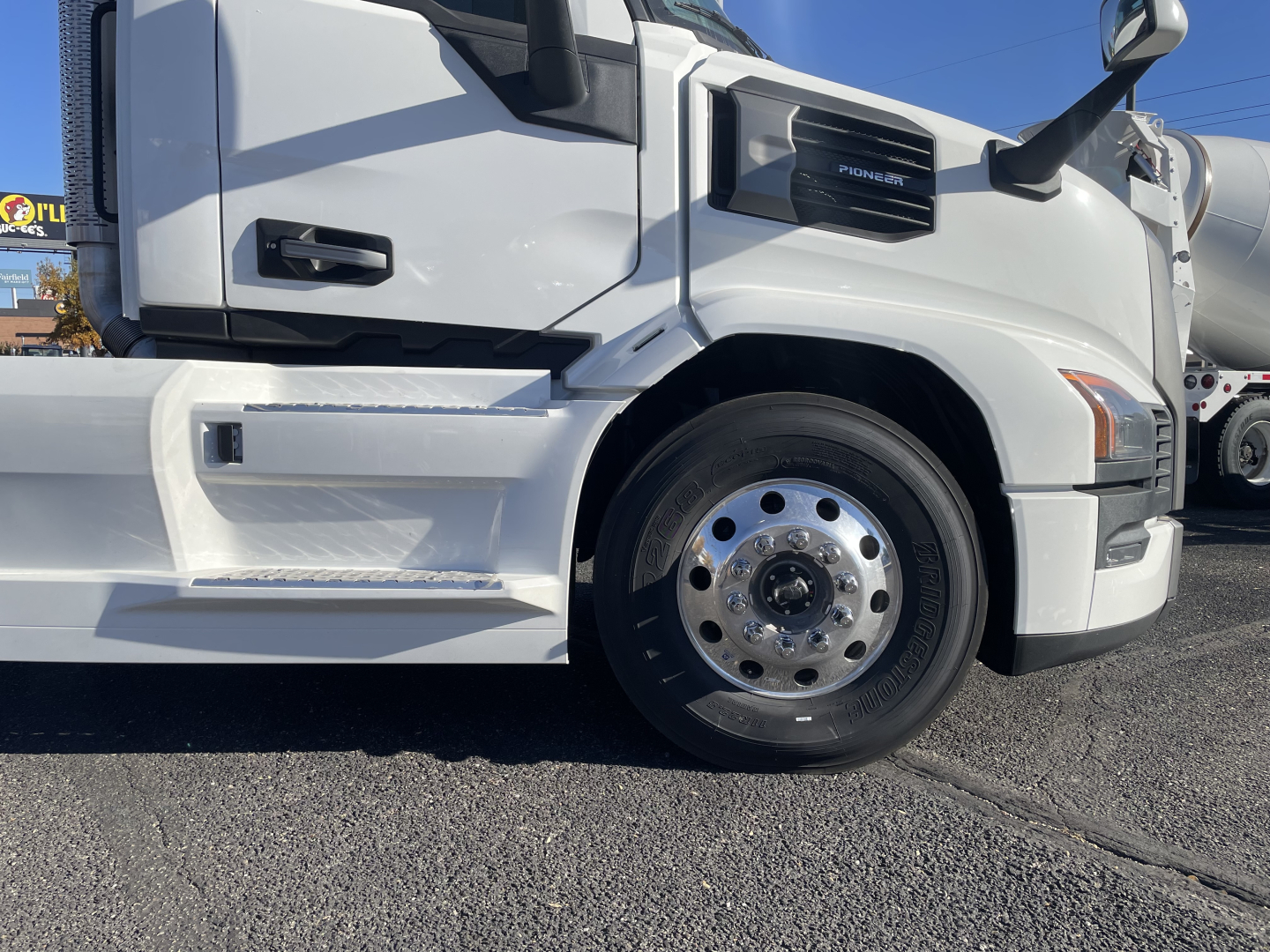 Front half of a white Mack PR64T truck showing front wheel, fender, and cab with step; visible badge and side mirror.