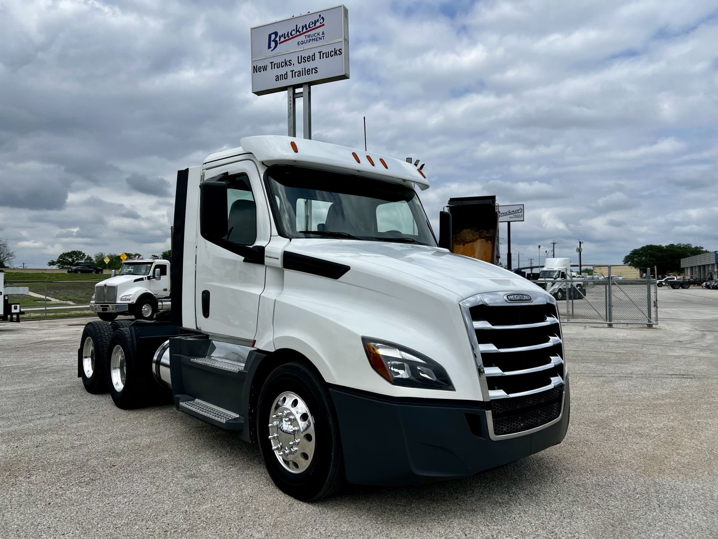 Freightliner Cascadia 126 daycab tractor in a dealership lot, white cab, chrome grille, side mirrors, with a sign in background and a trailer behind.