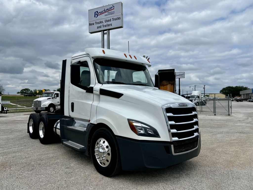 Freightliner Cascadia 126 daycab tractor in a dealership lot, white cab, chrome grille, side mirrors, with a sign in background and a trailer behind.