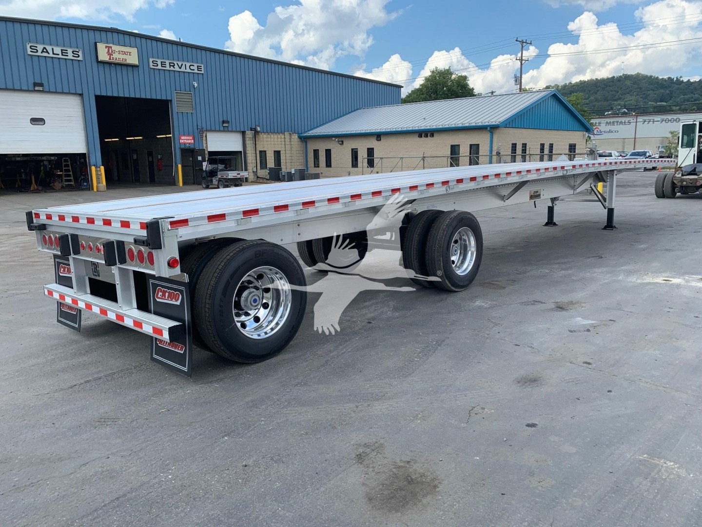 Flatbed trailer showing dual axles with chrome wheels and red/white reflective tape along the sides in a dealership lot.