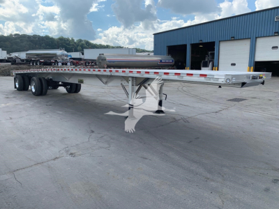 Flatbed trailer in a yard showing chrome beam with red/white reflective tape, dual rear axles, and landing gear in front; building in background.
