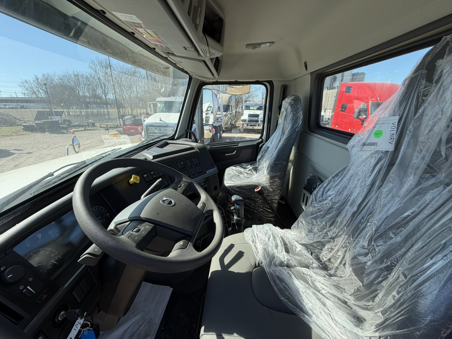 Cockpit view of a new Volvo VHD86B300 cab showing steering wheel, dashboard, and wrapped driver seat in plastic. Windows reveal outdoor yard with other trucks.