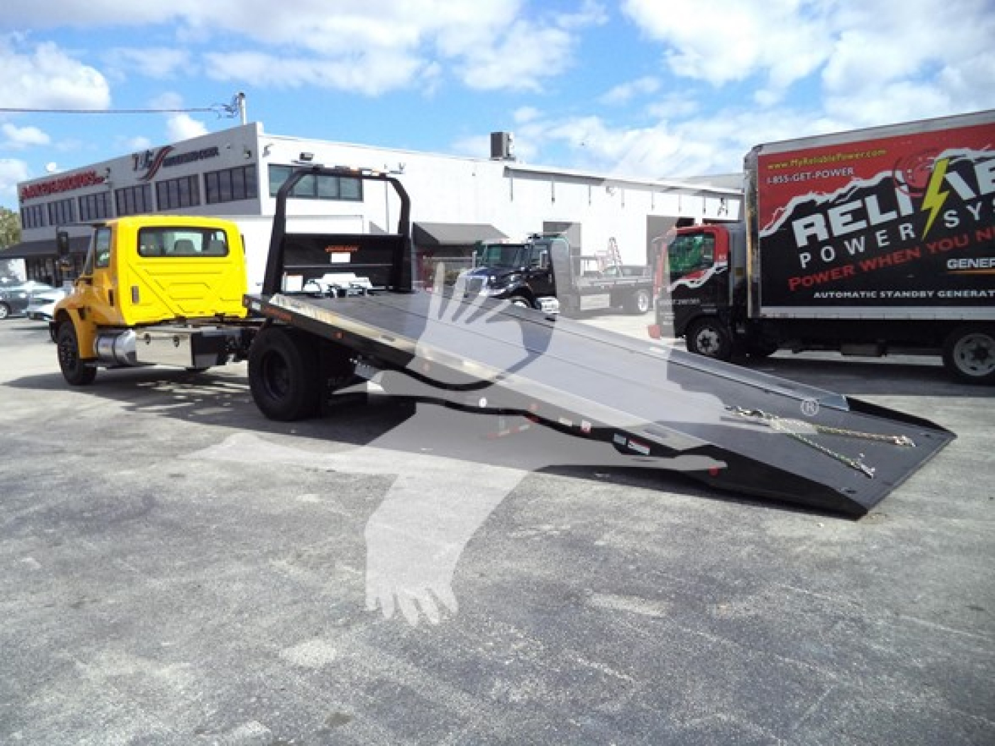 Truck bed with a yellow International Durastar tow truck and a flatbed rollback in a lot; other trucks in background.