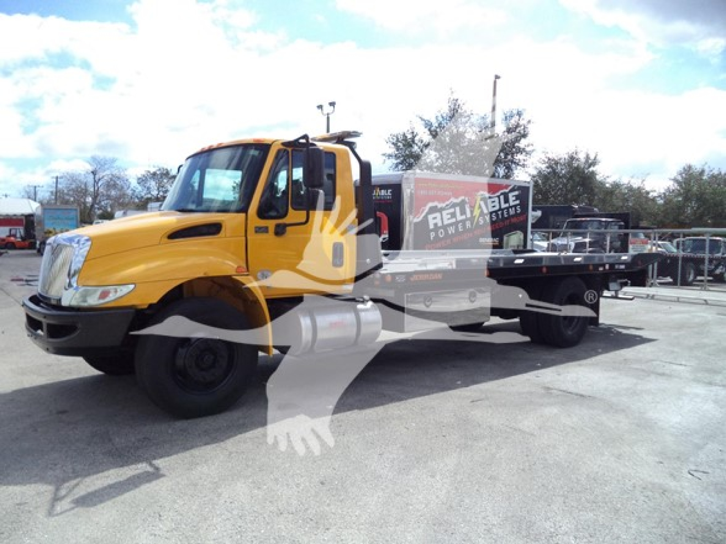 2019 International Durastar 4300 rollback tow truck in yellow with flatbed, side view showing cab and bed with 'Reliable Power Systems' trailer inside lot.
