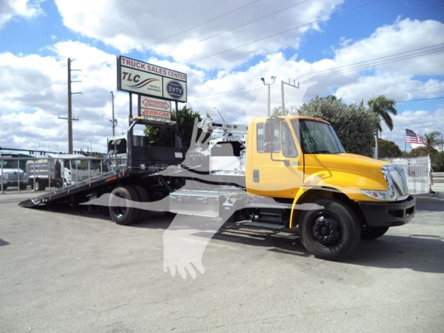 Tow truck on a rollback bed with a yellow cab; side view shows metal ramp bed and 'TRUCK SALES CENTER' sign in background.