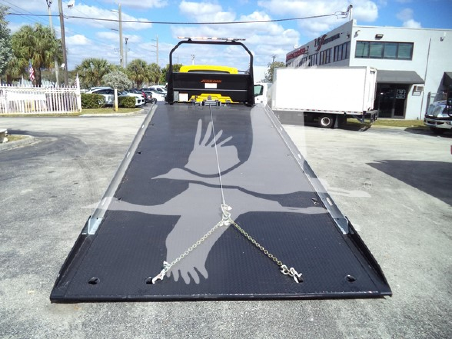 Flatbed tow truck bed with a chain tie-downs, rear of cab behind, and a crane in the distance. No legible labels visible on bed surface.