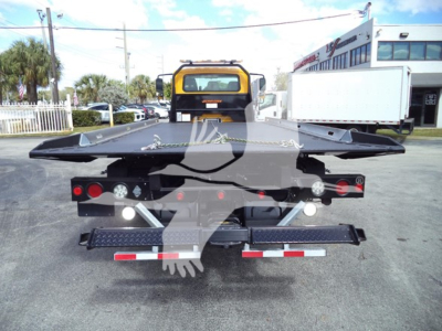 Back of rollback tow truck with flatbed and black chain hooks, taillights, and rear bumper; yellow crane/cab in distance.