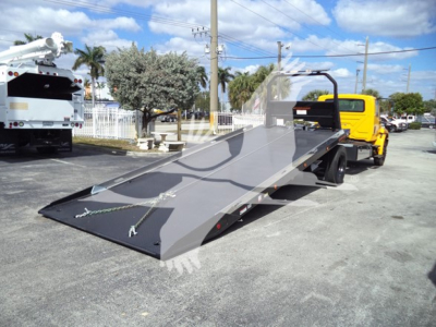 Truck in a lot with a flatbed rollback body showing a lowered steel ramp and deck. Side panels and hydraulic cradle visible.