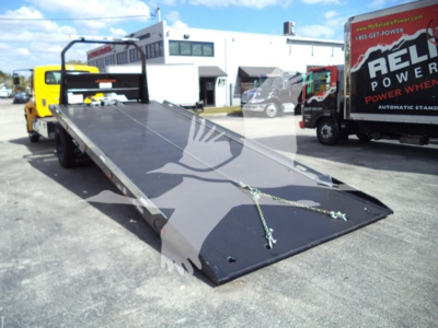 Flatbed tow truck bed with hydraulic plate in lowered position; black steel surface with chains, rear ramps raised; service bay in background.