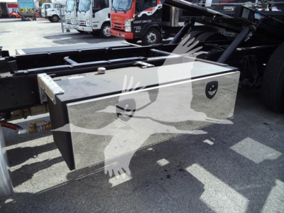 Truck chassis with a large metal toolbox mounted between the frame rails of a rollback tow truck; several other trucks in the background.