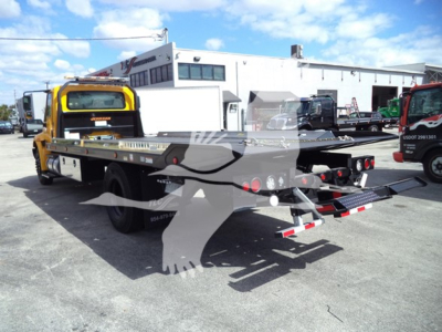 Flatbed rollback tow truck rear view showing hydraulic ramps extended and warning lights; background shows a dealership lot with multiple trucks.
