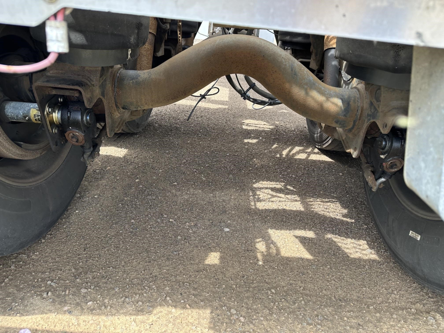 Underneath a trailer showing axles, suspension, and a curved exhaust/air line between wheels; view focuses on the undercarriage and piping.