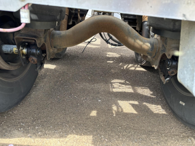 Underneath a trailer showing axles, suspension, and a curved exhaust/air line between wheels; view focuses on the undercarriage and piping.
