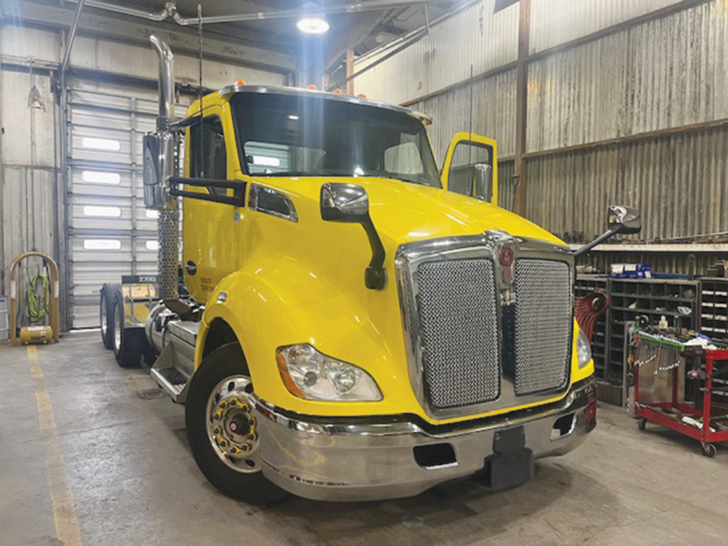 Kenworth T680 day cab truck in a workshop showing bright yellow cab and chrome grille with large front bumper.