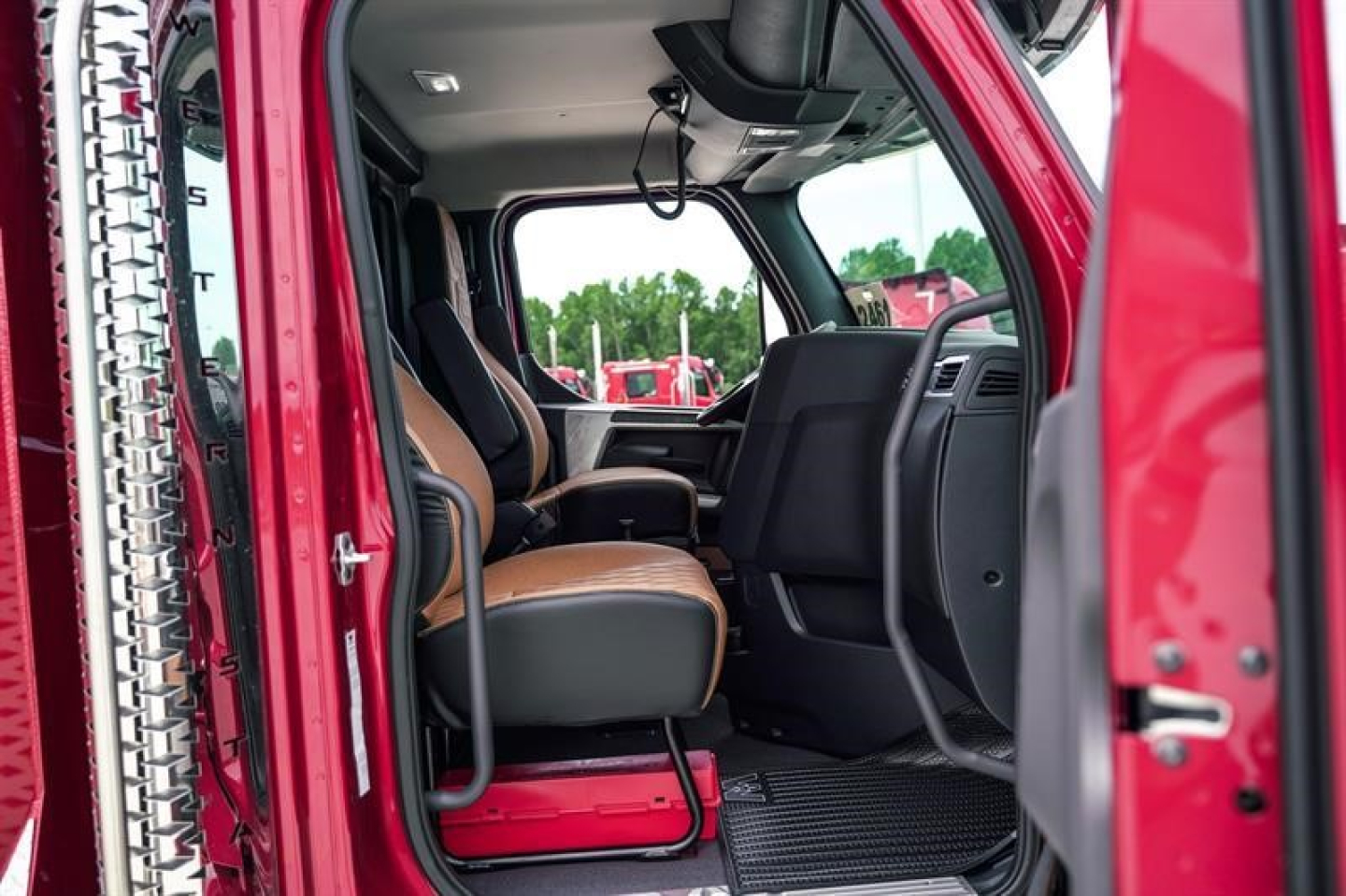 Truck cabin interior of a Western Star 47X with red exterior, front bench seat, open door, view of driver and passenger seats from rear door, dashboard visible.