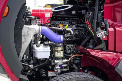 Truck engine bay of a 2027 Western Star 47X showing coolant reservoir, blue intake hose, various hoses, and components.