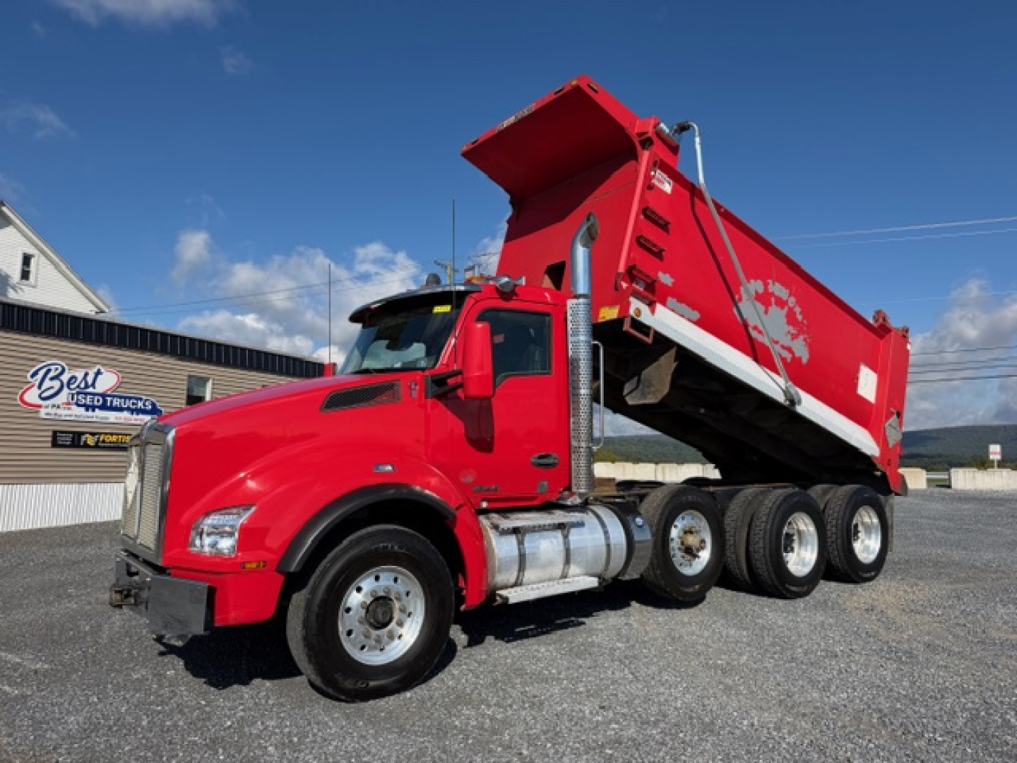 Kenworth T880 dump truck in red with bed raised showing hydraulic components and triple rear axles.