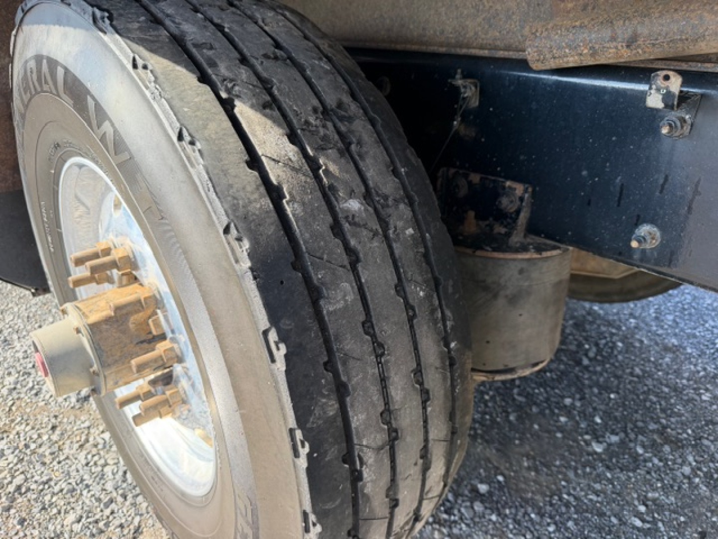 Kenworth T880 wheel and dual rear tire view showing tire tread and wheel hub with gold lug nuts; part of chassis and undercarriage visible.