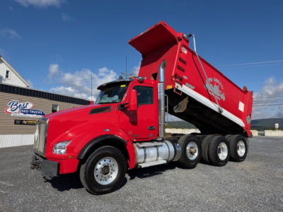 Kenworth T880 dump truck in red with bed raised showing hydraulic components and triple rear axles.