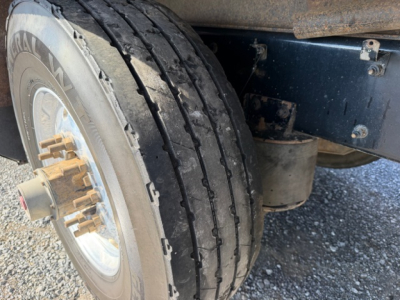 Kenworth T880 wheel and dual rear tire view showing tire tread and wheel hub with gold lug nuts; part of chassis and undercarriage visible.
