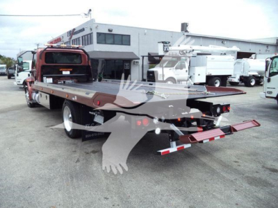 Flatbed tow truck in a dealership yard showing rear view with extended bed and reflective safety decals.