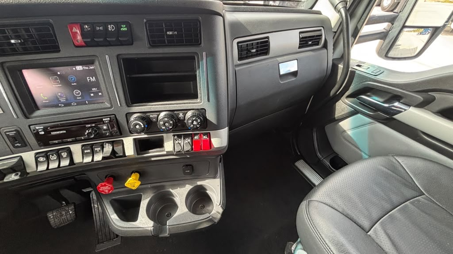 Truck cab interior showing driver seat and dashboard of 2023 Kenworth T680 with center console controls and cup holders.
