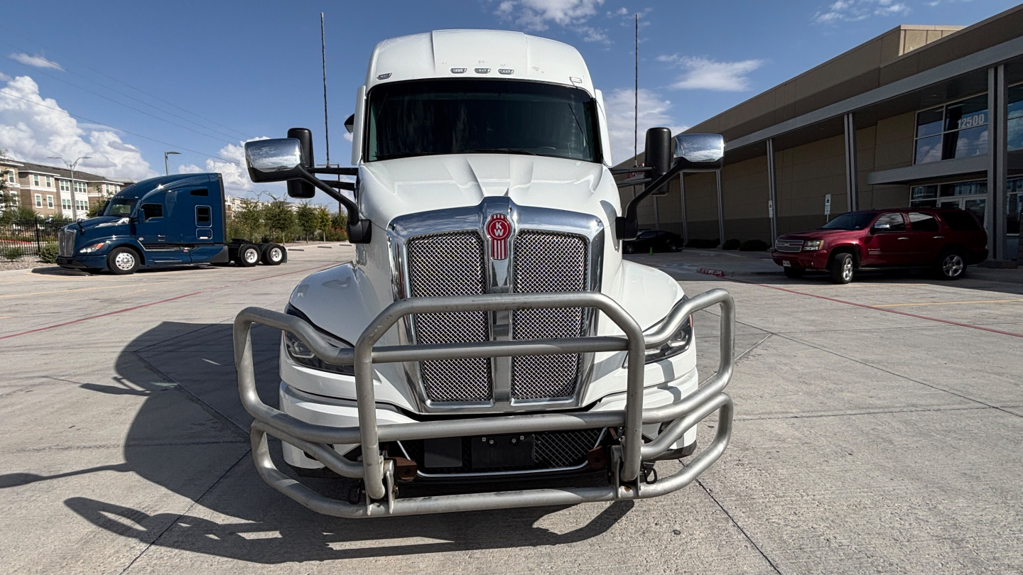 Kenworth T680 conventional sleeper truck front view with grille guard and chrome bumper, white paint, parking lot backdrop.