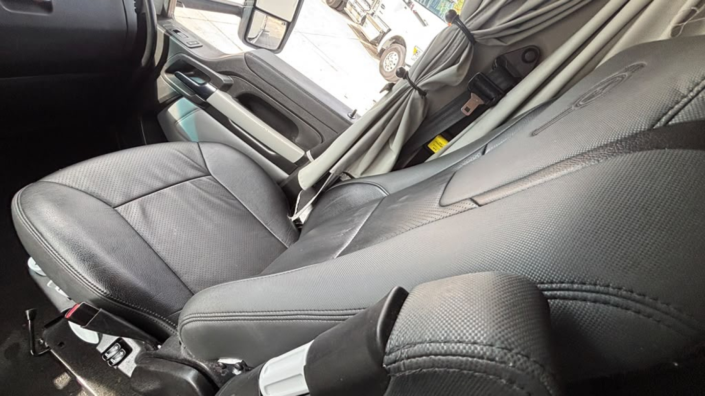 Interior of a Kenworth truck cab showing black leather seat and steering wheel area with window controls and seat belt.