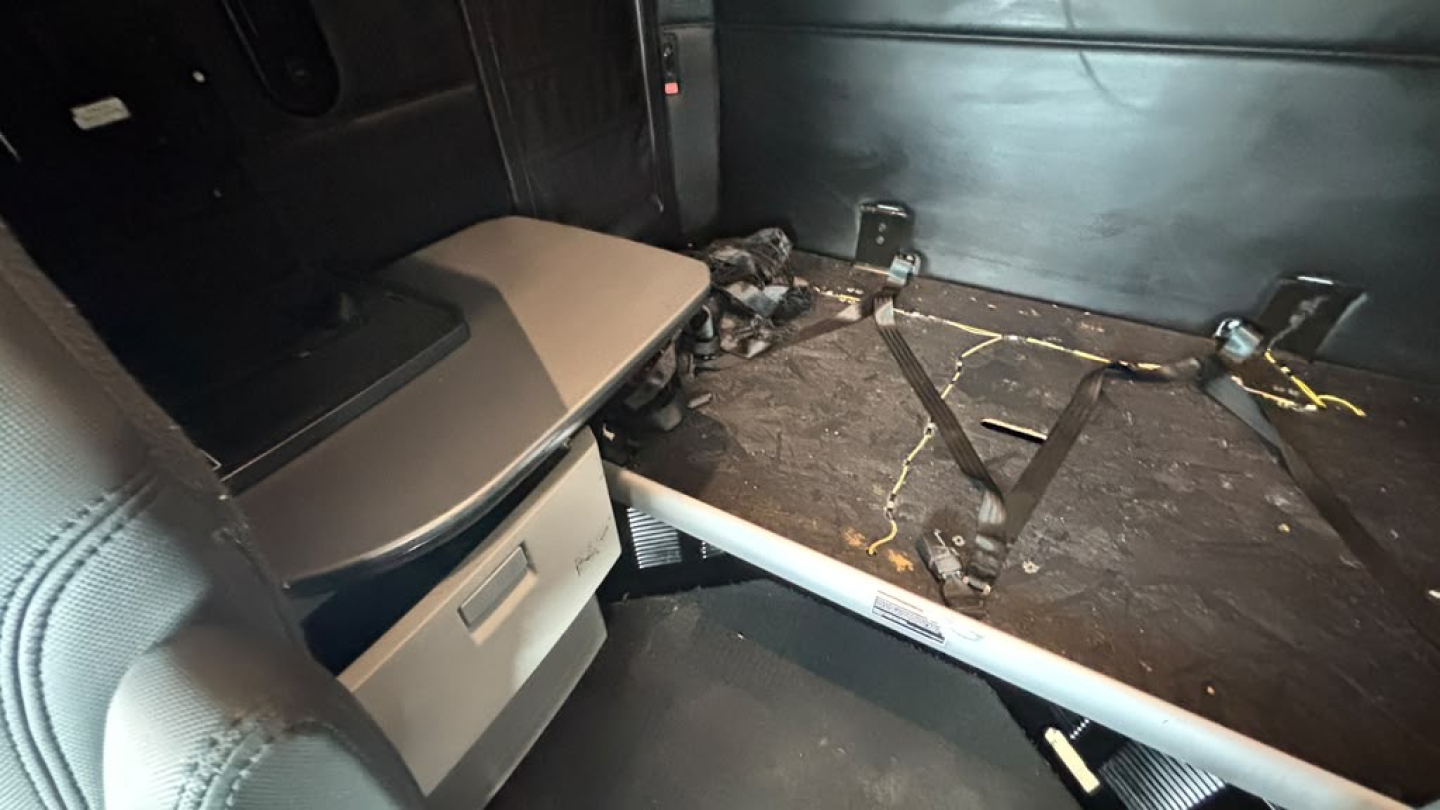 Truck interior cargo area of a Kenworth T680 showing a storage drawer under the sleeper partition and a flat floor with some debris and a seat edge.