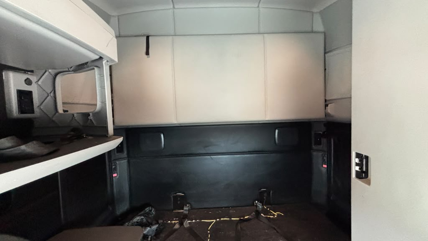 Truck cab interior showing sleeper bunk area with overhead cabinet and side shelf; features a light gray upper panel, black lower panel, and wiring on the floor.