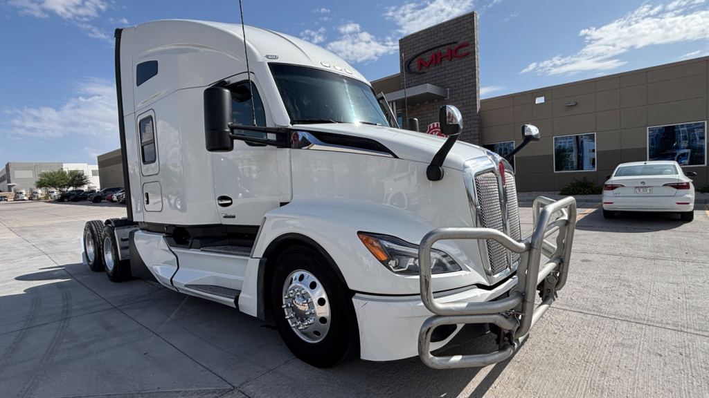 Kenworth T680 tractor-trailer in white, front angle view with chrome grille guard, parked outside showroom.