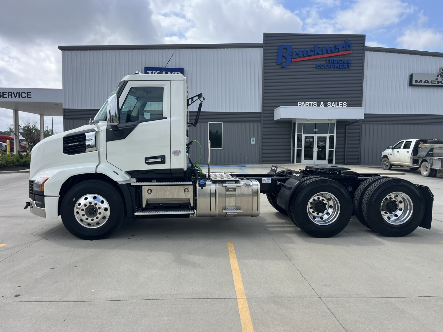 Truck chassis side view showing 2027 Mack AN64T cab-over-engine with white exterior, fuel tanks, and dual rear axles in a dealership lot.