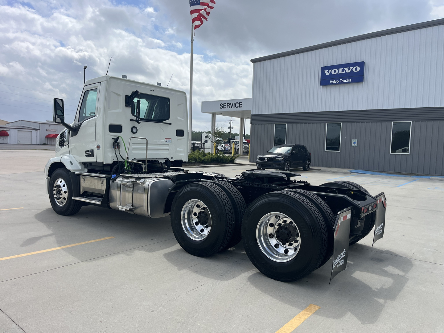 Truck chassis with dual rear axles and white Mack AN64T cab in a dealership lot, service building in background, VIN and stock visible.