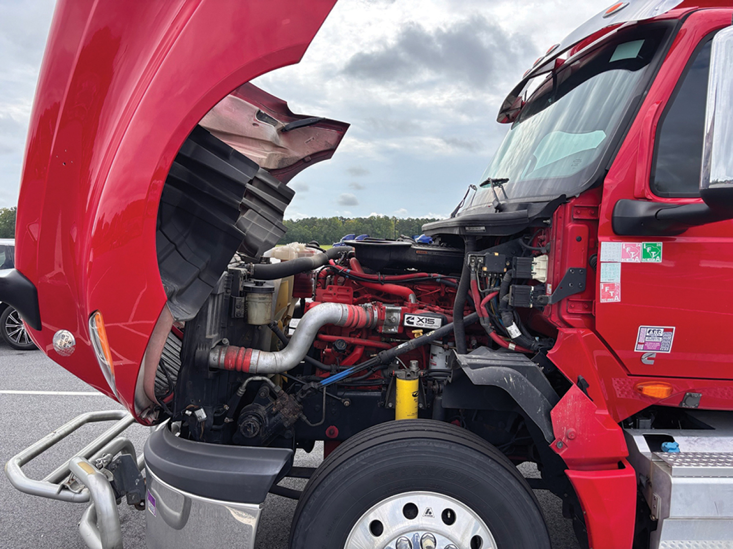 Truck engine bay of a 2020 International LT625 with red cab open hood showing engine components, hoses, and headlights; front left wheel visible.