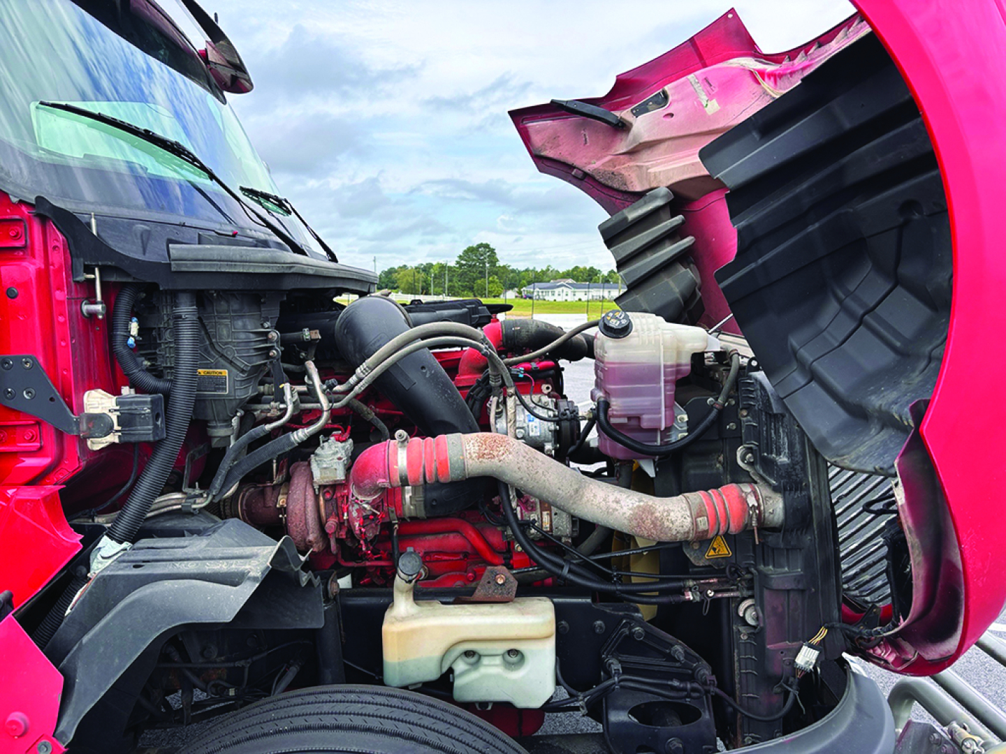 Engine bay of a red/pink 2020 International LT625 truck showing radiator, hoses, coolant reservoir, and various engine components.