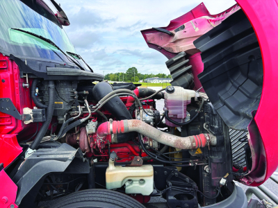 Engine bay of a red/pink 2020 International LT625 truck showing radiator, hoses, coolant reservoir, and various engine components.