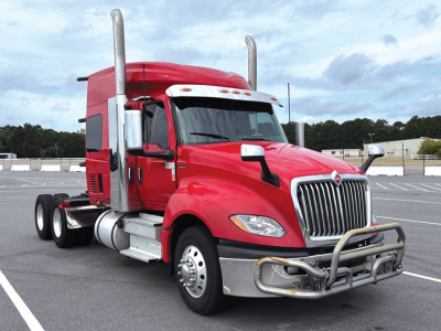 Truck cab with red 2020 International LT625 sleeper in a parking lot, showing front grille, bumper guard, chrome exhaust stacks, and dual rear axles.