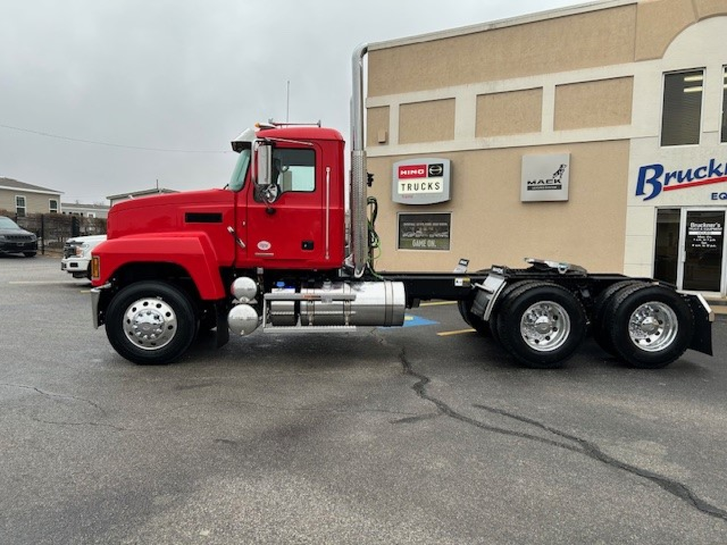 Mack PI64T truck cab and chassis in red showing side view with fuel tanks and dual rear axles; dealership building in background.