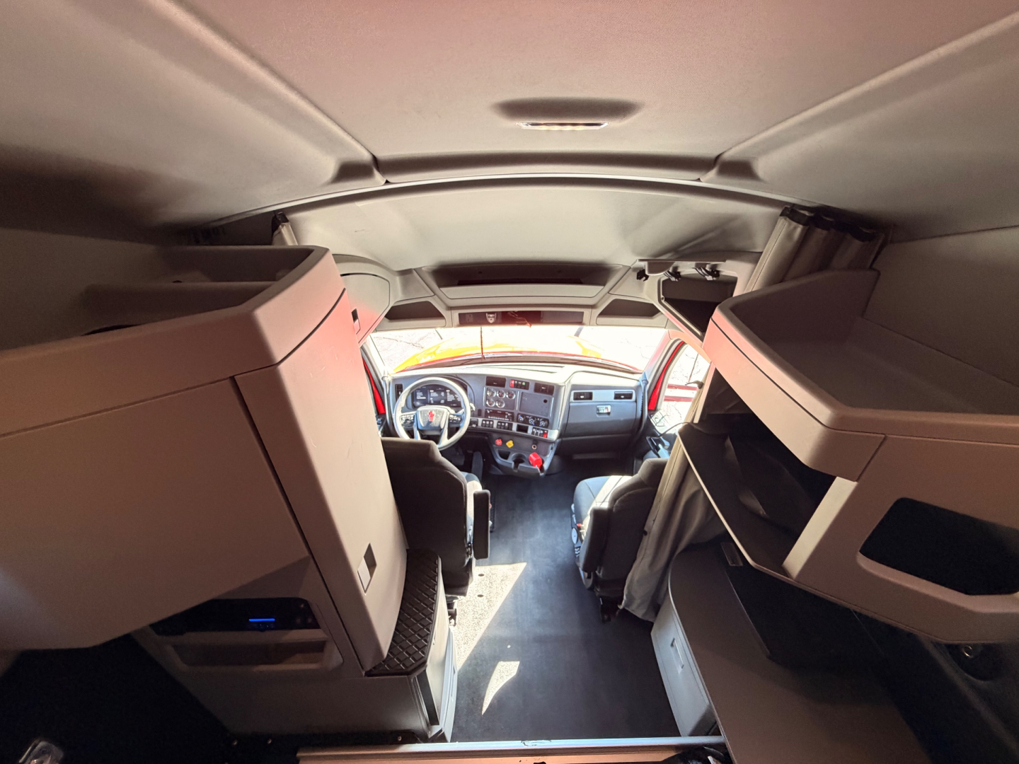 Kenworth T680 truck cab interior view from rear looking toward the dash, with sleeper area on both sides and overhead storage compartments.